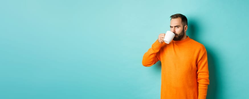 Image of guy drinking coffee and looking suspicious at camera, stare at something strange, standing in orange sweater over turquoise background