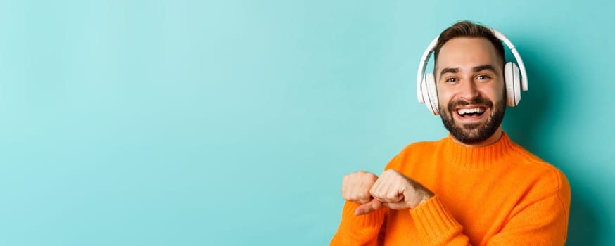 Close-up of handsome modern man listening music in headphones, standing in orange sweater over turquoise background