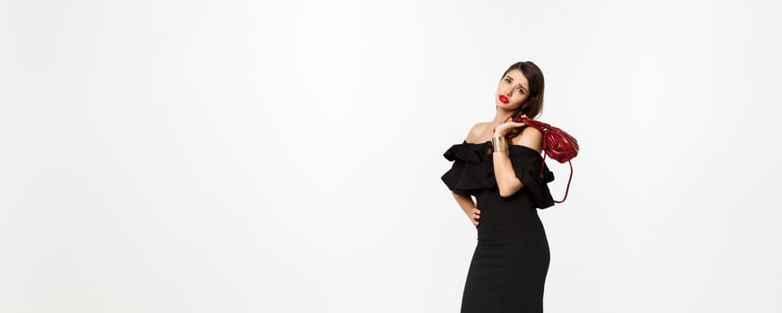 Beauty and fashion concept. Full length of tired young woman in high heels and elegant dress, holding purse on shoulder and looking with fatigue at camera, white background.