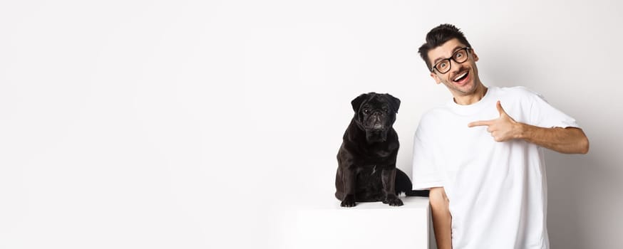 Happy young man showing his cute dog, pointing finger at black pug and smiling, standing over white background