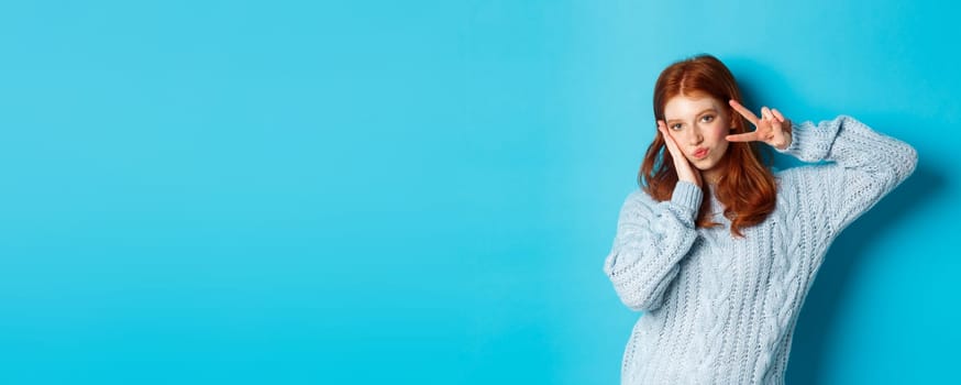 Modern teen girl with red hair, showing peace sign and posing in sweater against blue background
