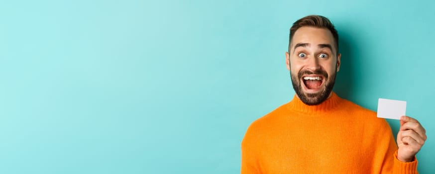 Close-up of excited caucasian man showing his credit card, smiling and staring amazed, standing in orange sweater against turquoise background