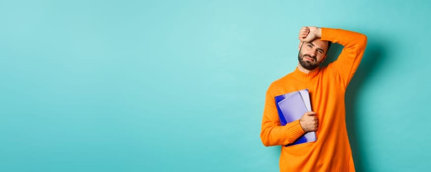 Education. Tired male student holding notebooks and wiping sweat off forehead, looking exhausted, standing in orange sweater against turquoise background