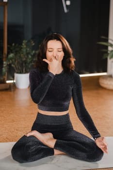 A woman in a black tracksuit sits on a yoga mat in a lotus position in the gym