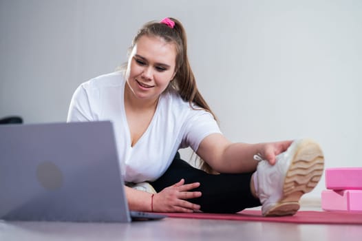 Young caucasian fat woman doing bends on a sports mat and watching a training video on a laptop. A chubby girl doing stretching remotely using video communication.