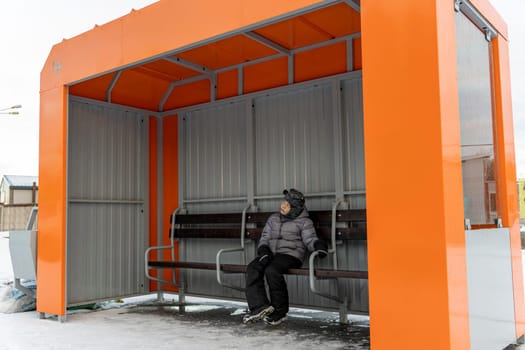 a lonely child waiting for a bus at an empty bus stop in winter. A small orange bus stop and a waiting boy sitting on a bench on a cold winter day.