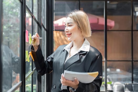 girl asian woman entrepreneur of small company putting a adhesive sticky notes in glass wall in office during analyzing formulating business strategies.
