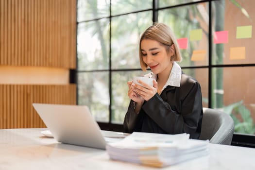 Smiling business woman drinking coffee while working on laptop from office
