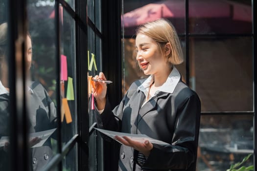 girl asian woman entrepreneur of small company putting a adhesive sticky notes in glass wall in office during analyzing formulating business strategies