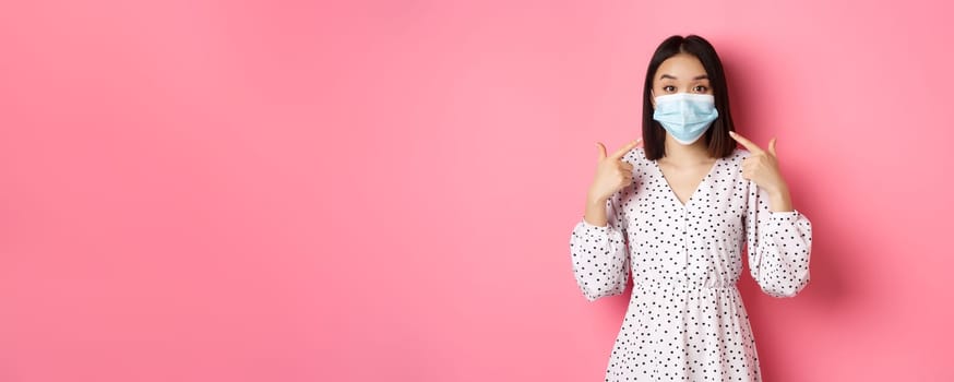 Coronavirus, social distancing and lifestyle concept. Cute asian woman pointing at face mask, asking to use measures against covid-19, standing over pink background
