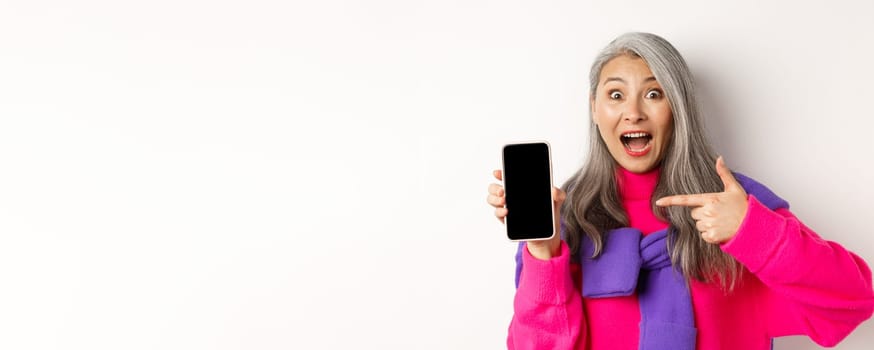 Online shopping. Beautiful asian grandmother smiling, pointing finger at smartphone blank screen, looking amazed, showing mobile application, standing over white background