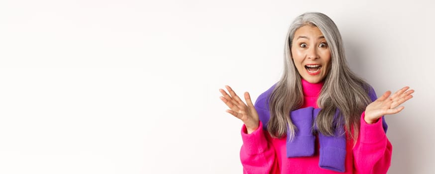 Close-up of excited asian senior woman spread hands sideways, gasping and looking amazed and surprised, hear great news, standing over white background