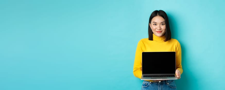Beautiful and stylish asian woman demonstrate product on screen, showing empty laptop display and smiling, standing over blue background