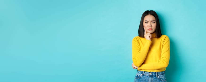 Image of pensive and serious asian woman touching chin, frowning and staring at camera puzzled, making difficult choice, standing over blue background