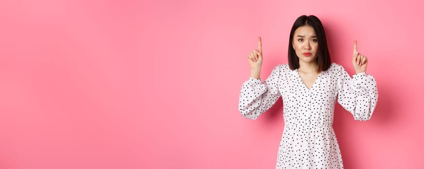 Upset asian girl pointing fingers up, showing promo banner, staring at camera with disappointed gloomy look, standing in dress over pink background
