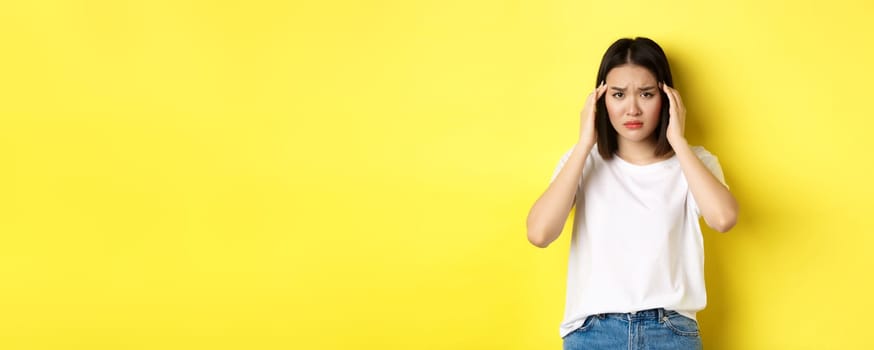 Sad asian girl touching head and frowning, feeling sick, having headache, standing in white t-shirt over yellow background