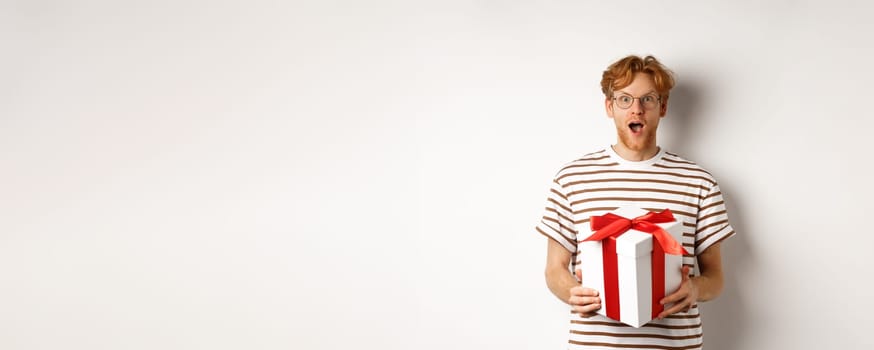 Valentines day and holidays concept. Surprised redhead boyfriend in glasses looking grateful at camera, receive big present in box, standing over white background