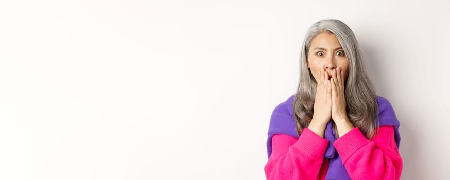 Close up of shocked asian old woman with grey hair, standing in stylish pink sweater, gasping startled and amazed, staring at camera, white background