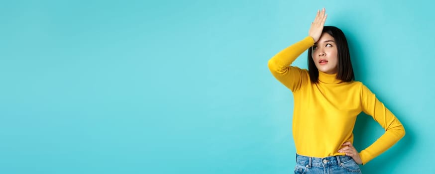 Emotions and lifestyle concept. Annoyed asian girl roll eyes and face palm, standing bothered in yellow pullover against blue background