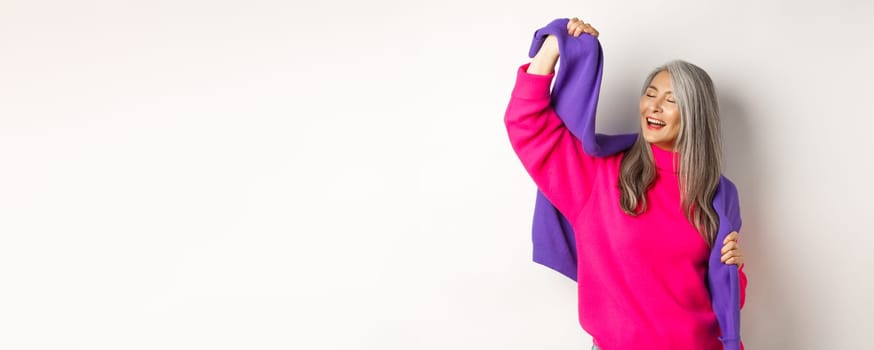 Carefree korean elderly woman in pink sweater, dancing with sweatshirt on shoulders and smiling, posing happy over white background