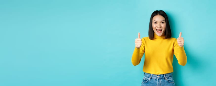 Beautiful asian woman praise good work, showing thumbs up gesture and smiling in approval, recommend product, standing satisfied over blue background