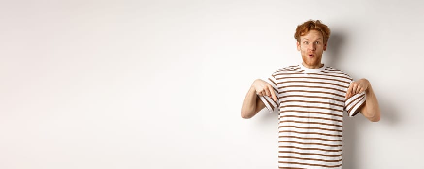 Image of funny redhead male student pointing fingers down, showing promo offer with excited smile, standing over white background