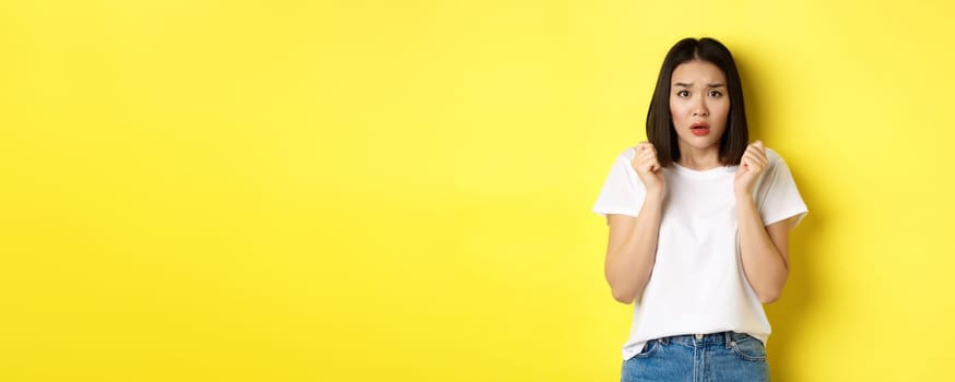 Beauty and fashion concept. Scared timid asian girl looking anxious, gasping startled and standing over yellow background