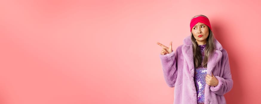 Fashion and shopping concept. Skeptical middle-aged asian woman in purple fur coat looking and pointing finger left with displeased, sad expression, standing over pink background