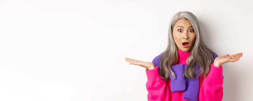 Close up of shocked asian mom staring at camera, spread hands sideways and gasping at camera confused, standing over white background