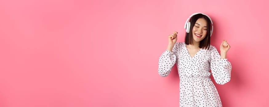 Happy young asian woman dancing and having fun, listening music in headphones, standing over pink background