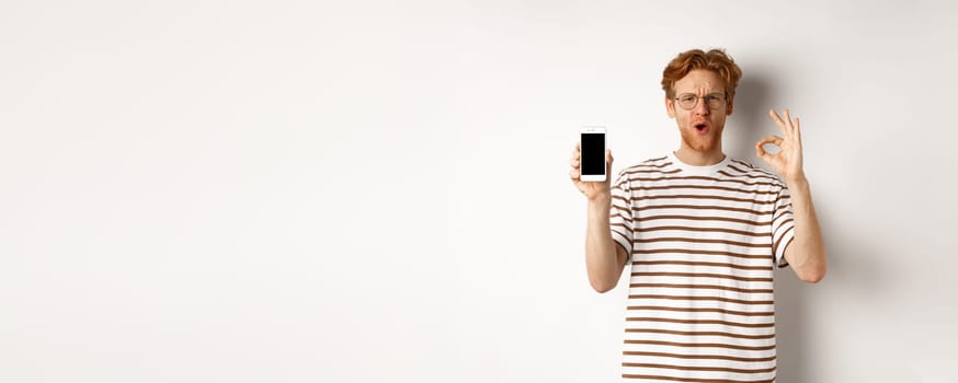 Technology and e-commerce concept. Young man with red hair showing okay sign and blank smartphone screen, praising awesome app or video game, white background.