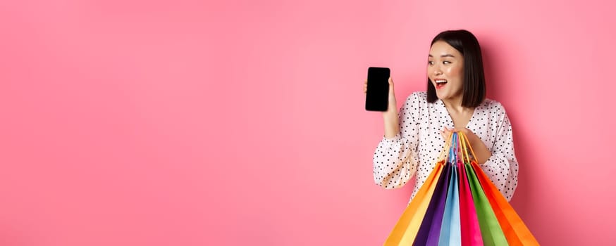 Attractive asian woman showing smartphone app and shopping bags, buying online via application, standing over pink background