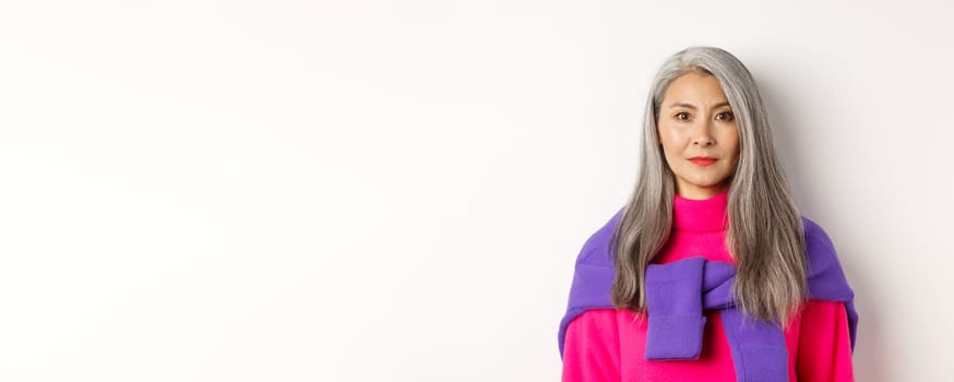 Close-up of stylish woman with grey hair and no wrinkles, smiling and looking at camera, standing in trendy outfit over white background