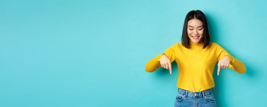 Shopping concept. Beautiful asian girl in yellow sweater smiling, pointing fingers down with satisfied face, showing cool advertisement, blue background