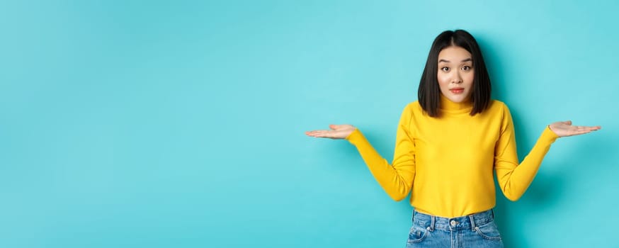 Image of indecisive asian woman shrugging shoulders, spread hands sideways and looking clueless at camera, standing indecisive against blue background
