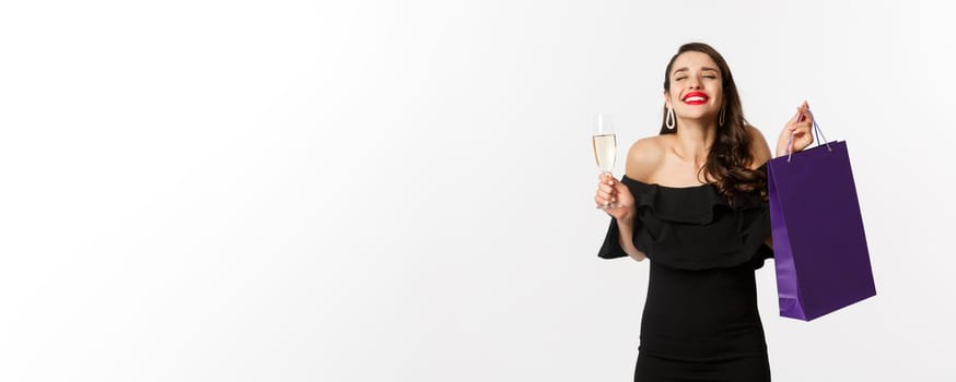 Happy smiling woman celebrating, holding present in shopping bag and glass of champagne, standing in black dress over white background