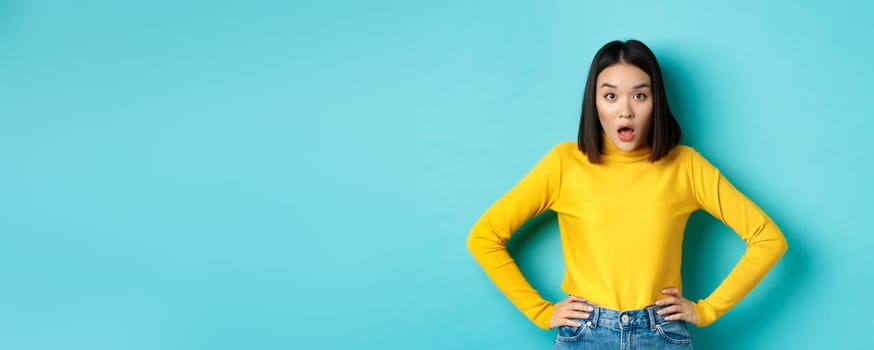 Portrait of surprised asian woman drop jaw, listening to big news, looking amazed at camera, standing in yellow sweater against blue background