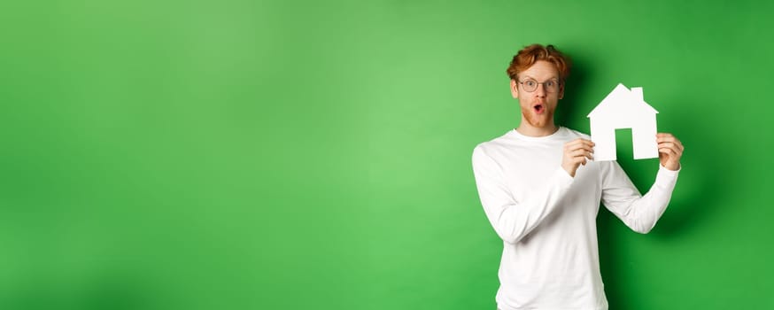 Real estate and buying property concept. Surprised young redhead man showing paper house model and looking amazed, standing over green background