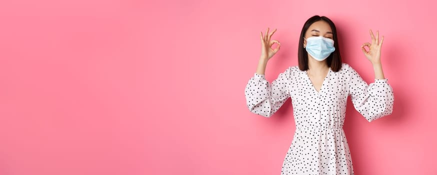 Coronavirus, social distancing and lifestyle concept. Satisfied asian woman in face mask showing okay signs, approve and agree, praising good choice, pink background