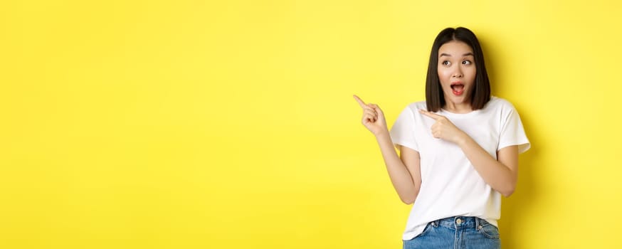 Beauty and fashion concept. Beautiful asian woman in white t-shirt pointing fingers left, standing over yellow background