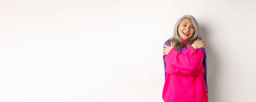 Valentines day and holidays concept. Lovely asian senior woman in pink sweater hugging herself with eyes closed, smiling, standing over white background