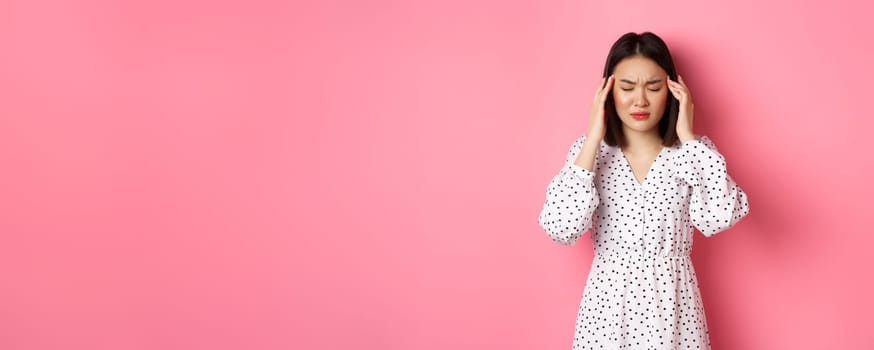 Image of upset asian woman having headache, feeling unwell or dizzy, close eyes and massaging head, suffering migraine, standing over pink background