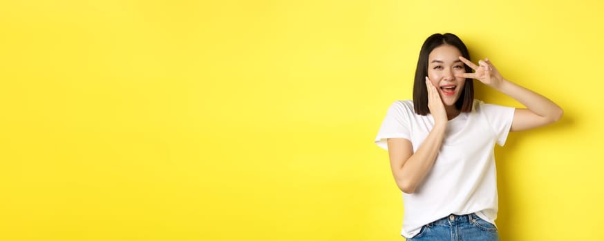 Lovely asian girl in white t-shirt posing with hand on cheek, showing peace sign on eye, standing over yellow background