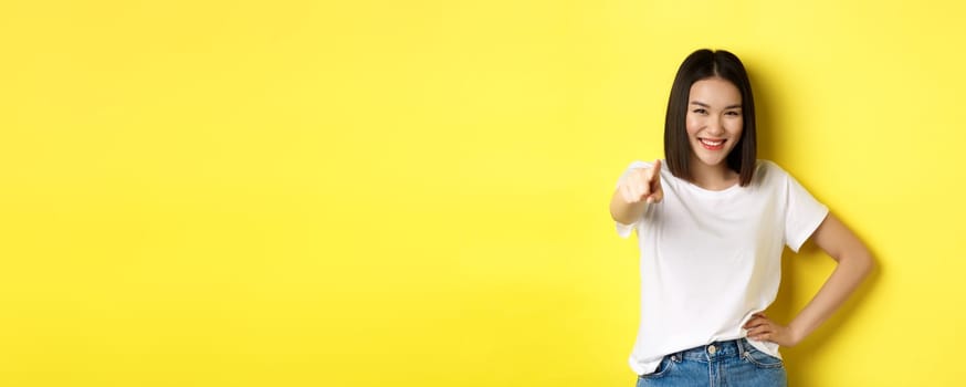 We need you. Confident asian woman smiling, pointing finger at camera, inviting to join her, beckon or choosing someone, standing over yellow background