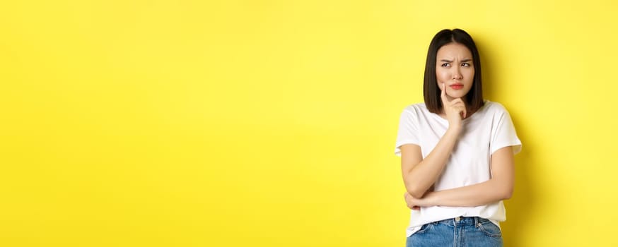 Beauty and fashion concept. Pensive asian woman thinking, looking thoughtful while pondering something, standing over yellow background