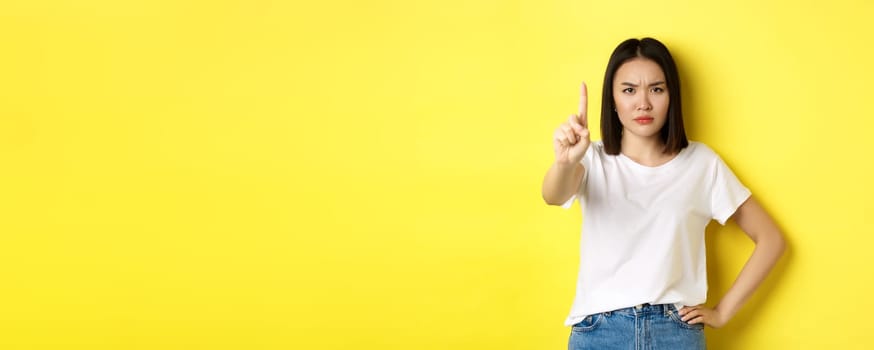 Confident and serious woman tell no, showing extended finger to stop and prohibit something bad, frowning and looking at camera self-assured, standing over yellow background