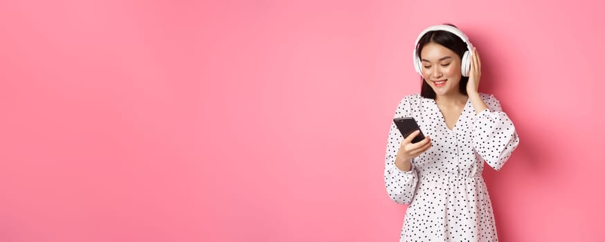 Cute asian girl listening music on headphones, looking at mobile phone and smiling, standing in dress against pink background