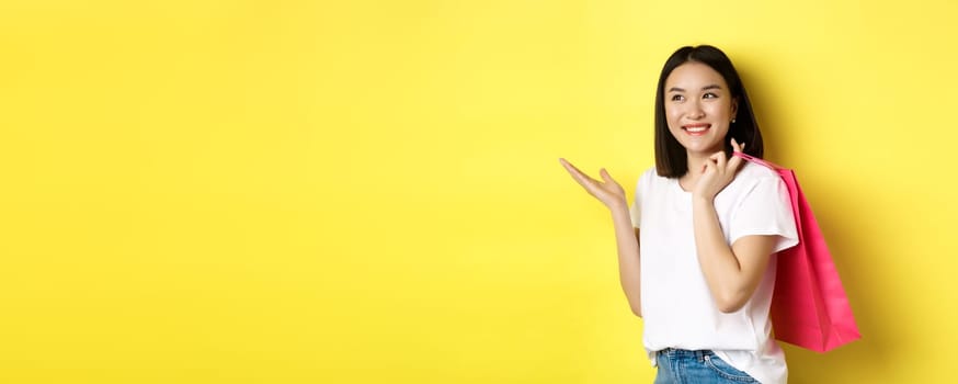 Cheerful asian female shopper looking amused, holding shopping bag and pointing hand left at store banner, standing over yellow background