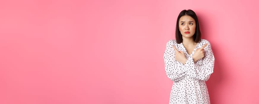 Concerned asian woman having doubts, pointing sideways and looking left with hesitant and sad face, need help with choice, standing over pink background