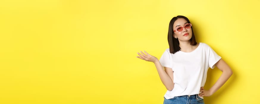 Fashion and lifestyle concept. Sassy and confident asian woman in trendy sunglasses looking self-assured at camera, standing over yellow background